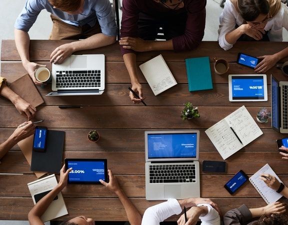 Group of people working around a wooden table with laptops, tablets, notebooks, and coffee cups.