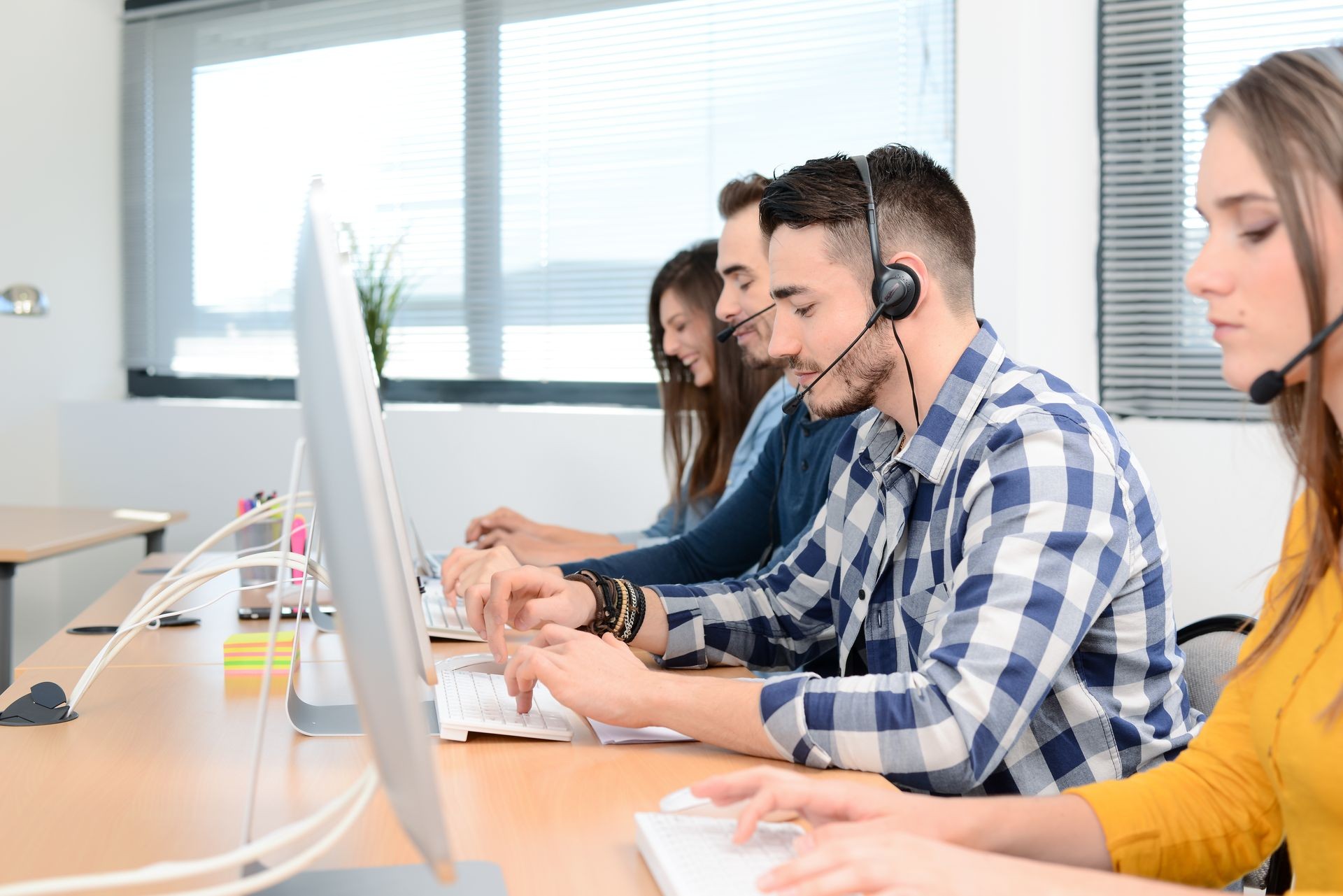 portrait of handsome and cheerful young man telephone operator with headset working on desktop computer in row in customer service call support helpline business center with teamworker in background